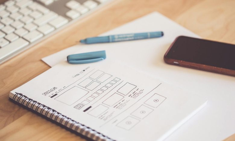 wireframing untuk pemula - A neat workspace featuring a sketchpad with wireframes, a smartphone, and a keyboard on a wooden desk.