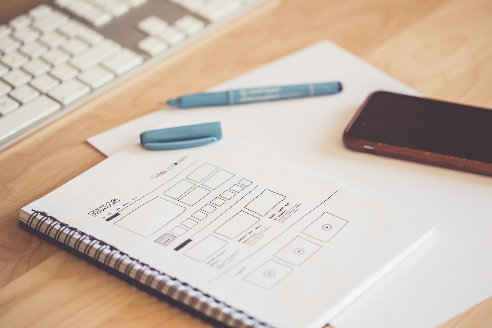 wireframing untuk pemula - A neat workspace featuring a sketchpad with wireframes, a smartphone, and a keyboard on a wooden desk.